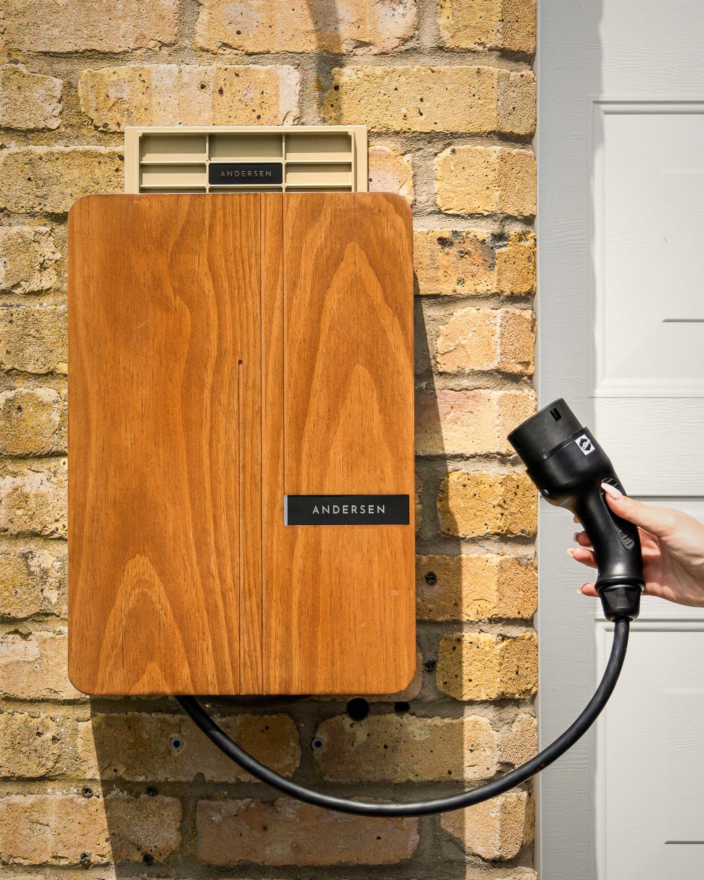 Borne de recharge murale en bois installée sur une façade en briques, avec câble de recharge à la main